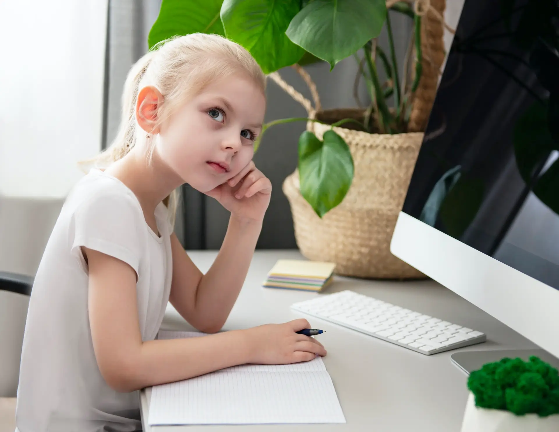 Young girl studying with notebook and computer at home desk with plant decoration.