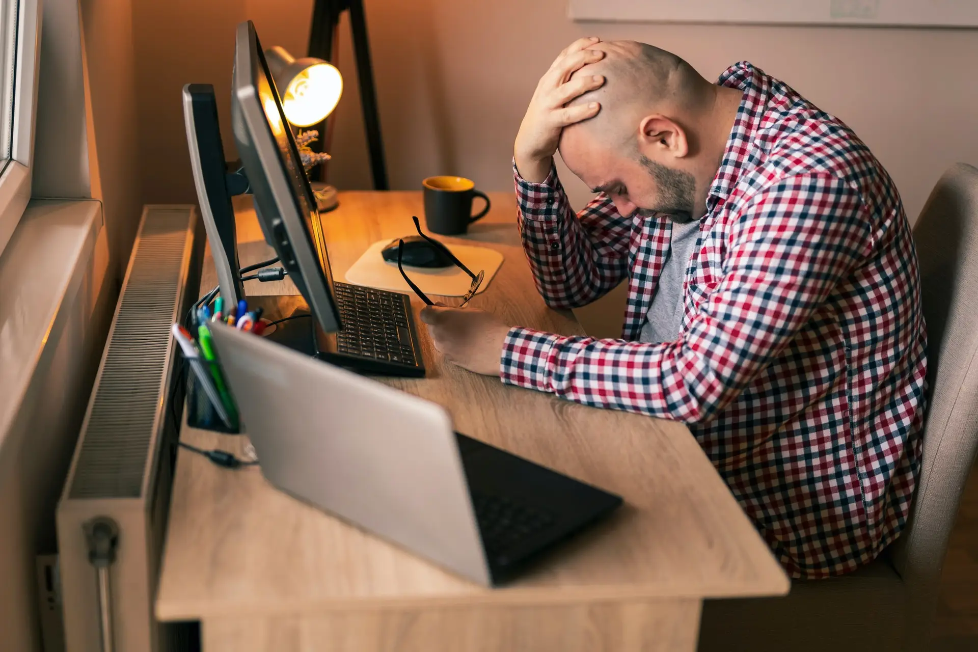 Stressed man at home office with head in hands facing computer and laptop on desk