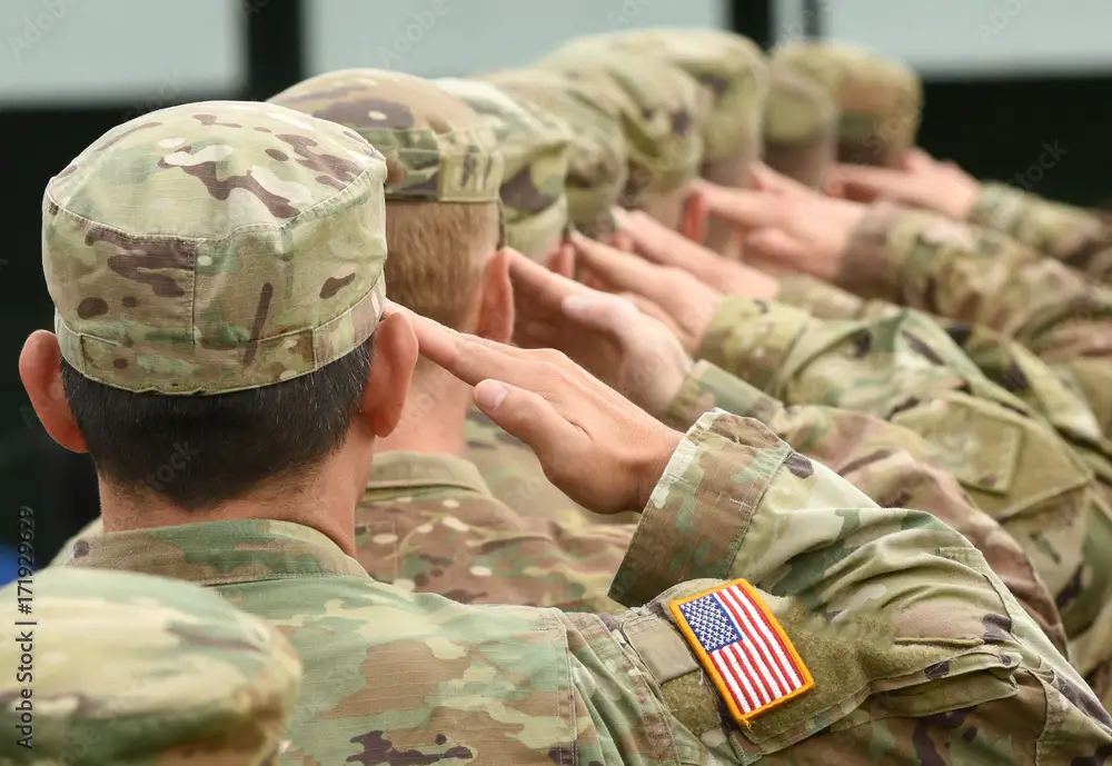 Group of military personnel saluting in camouflage uniforms with American flag patch on sleeves.