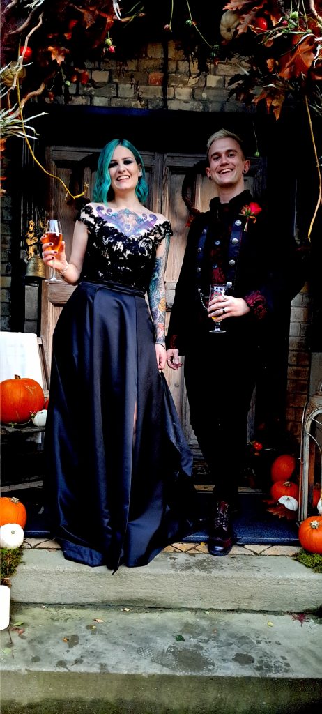 Pop-punk and emo wedding couple standing on gothic porch, dressed in alternative wedding attire, celebrating with drinks.