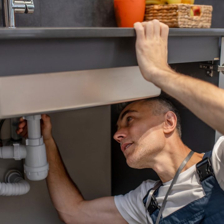 Man carefully examining bottom of sink and pipe Plumber, repair. Experienced attentive middle-aged man carefully examining bottom of kitchen sink touching pipe with his hand diagnosing problem