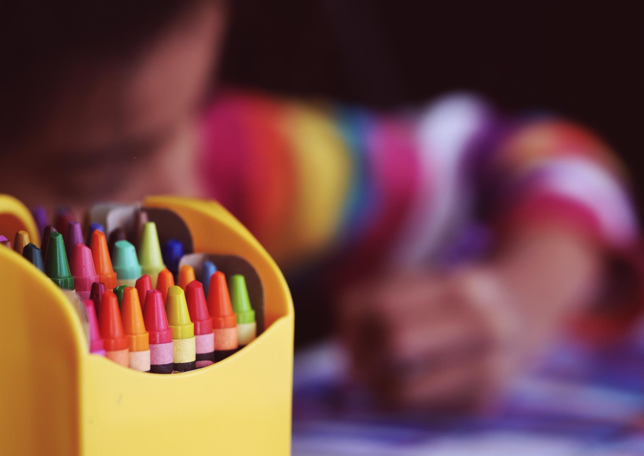 A child crayoning in the background, with a set of wax crayons in focus at the front of the picture