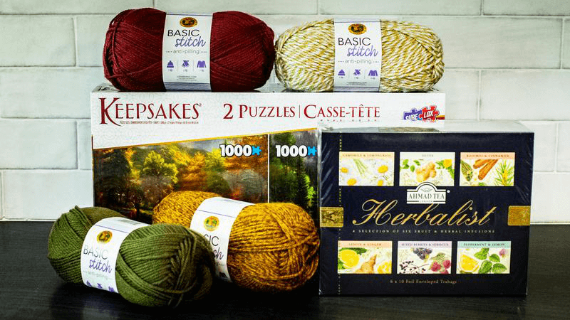 A picture of the Sip, Stitch, Solve gift set from Lionbrand Yarn. Pictured is the Herbalist one, which includes: a 2 puzzle box with nature scenario pictures, one summer and one in fall colors. There are also four skeins of Lionbrand yarn in fall colors, mustard yellow, deep green and red and a mouline yarn in white and golden yellow. At the front is a packet of herbalist tea with pictures of demon, ginger and a bounch of other herbs, berries and fruits on it. The Sip, Stitch, Solve gift sets bought from Lionbrand Yarn are perfect gifts when you want to show that you care. You can choose between Herbalist, Classical, or Fruity set.