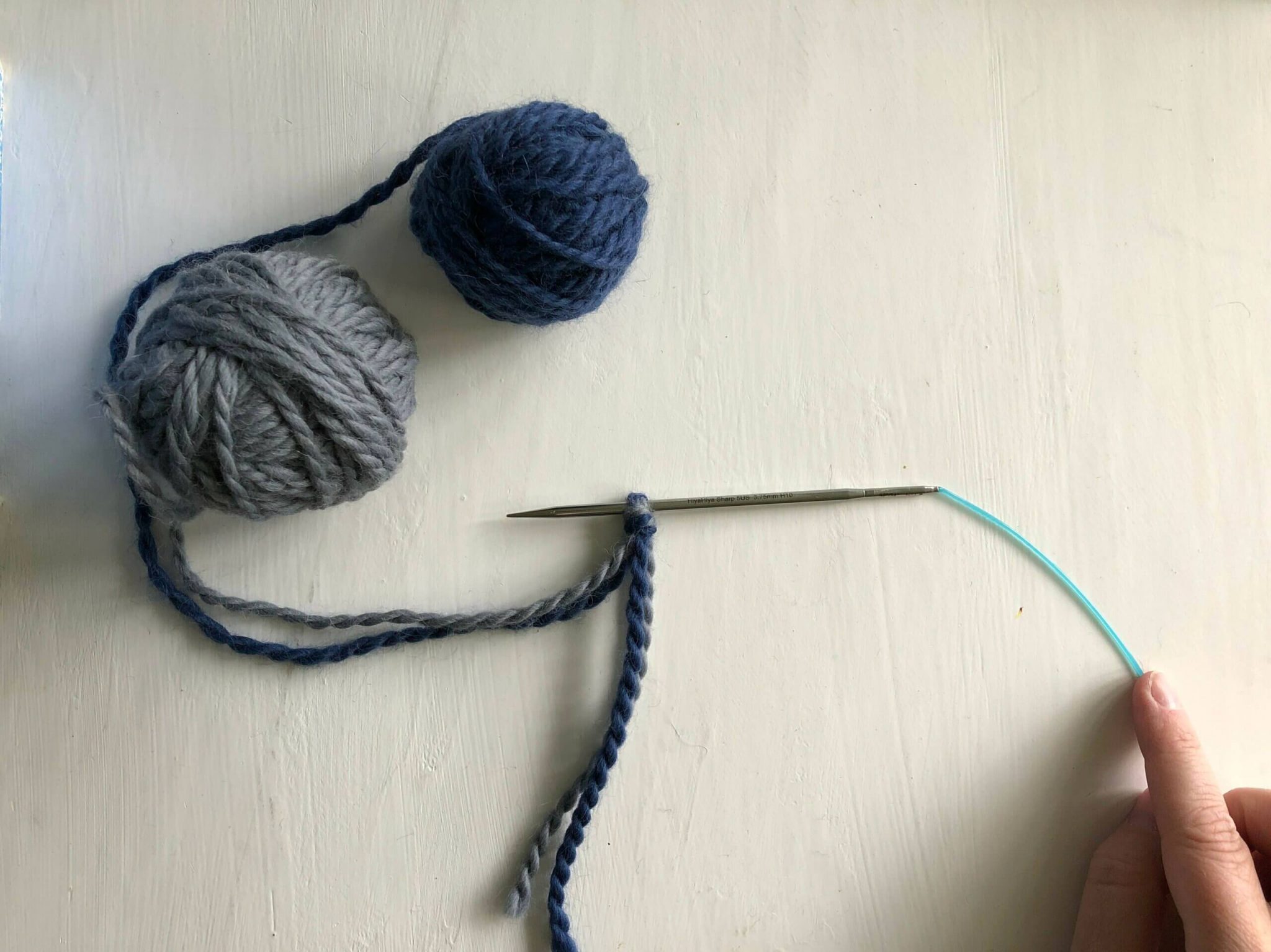 Pictures shows a knitting needle and a blue and a grey ball of wool yarn. The yarns are tied together in a slip knot that's attached to a knitting needle.