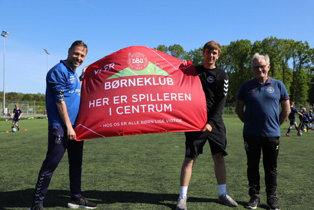 Slagelse B & I er blevet optaget som DBU Børneklub. Børneansvarlig Morten Weensgaard (tv) og børneudviklingstræner Kristian Vittenbach fik forleden overrakt et synligt bevis på den nye status af Henrik Oksholm (th) fra DBU Sjælland. Der er i øjeblikket 11 DBU Børneklubber på Sjælland. (Foto: Slagelse B&I.)