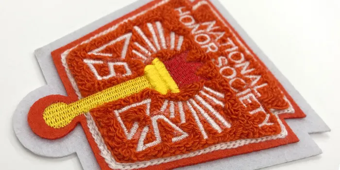 Orange chenillepatch med vit filtkant, broderad text och illustrerad gyllene fackla med röd flamma.