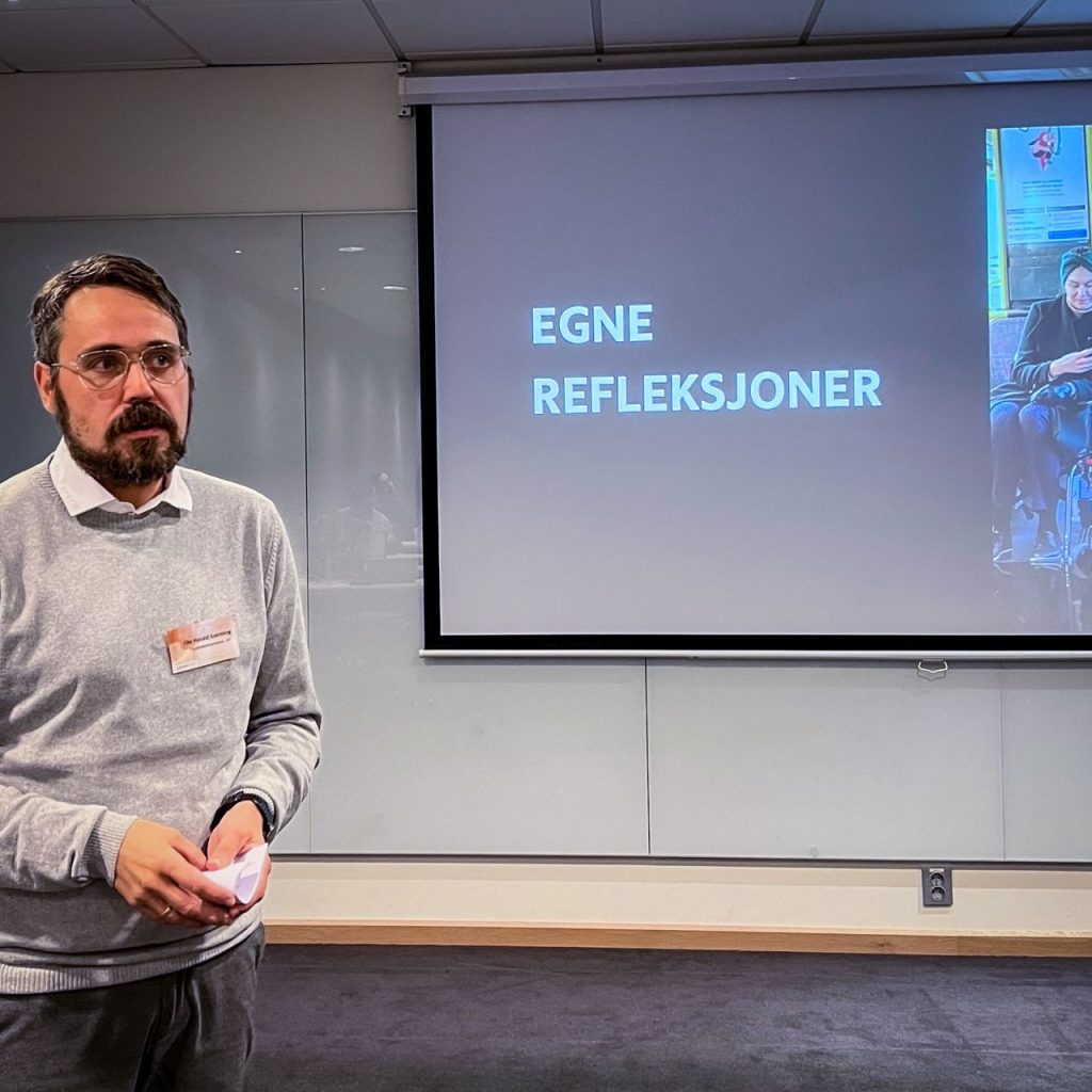 En mann med mørkt, kort hår, briller, skjegg og en lysegrå genser over en hvit skjorte står foran en powerpoint-presentasjon. Han ser ut mot publikum til høyre. På powerpointen står det "Egne refleksjoner" med store bokstaver ved siden av et bilde av en kvinne i rullestol.