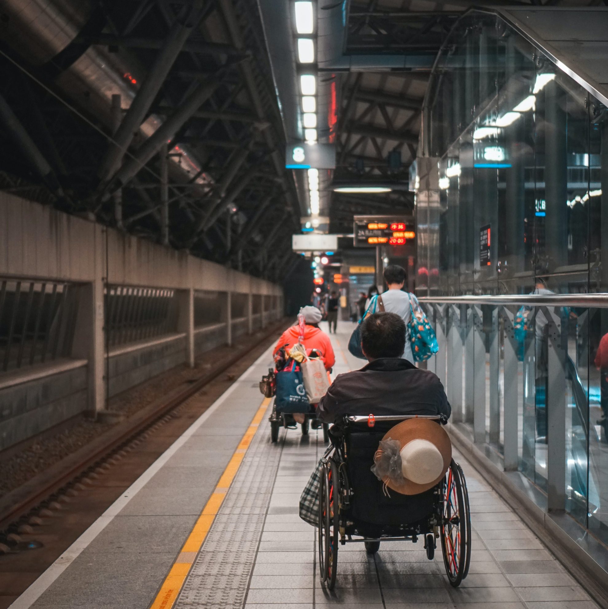 Togplattform med to personer i rullestol og én gående bakfra. I bakgrunnen skimtes asiatiske tegn på skiltene for togavganger. Fotografi.