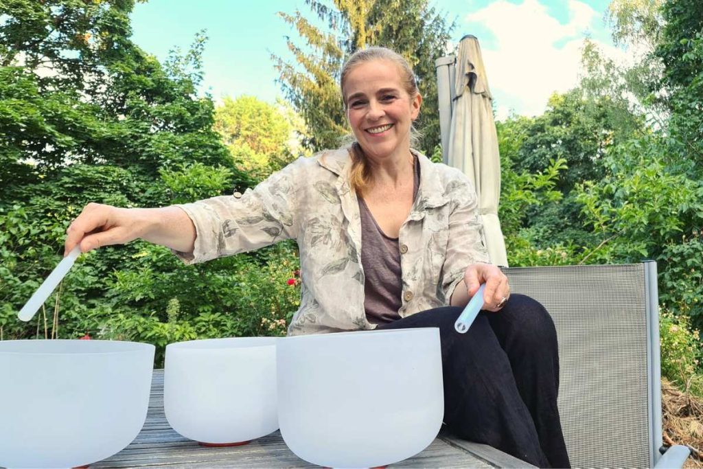 Elena Deppe spielt drei weiße Kristall-Klangschalen mit zwei Silkon-Reibern draußen am Gartentisch bei Sonne.