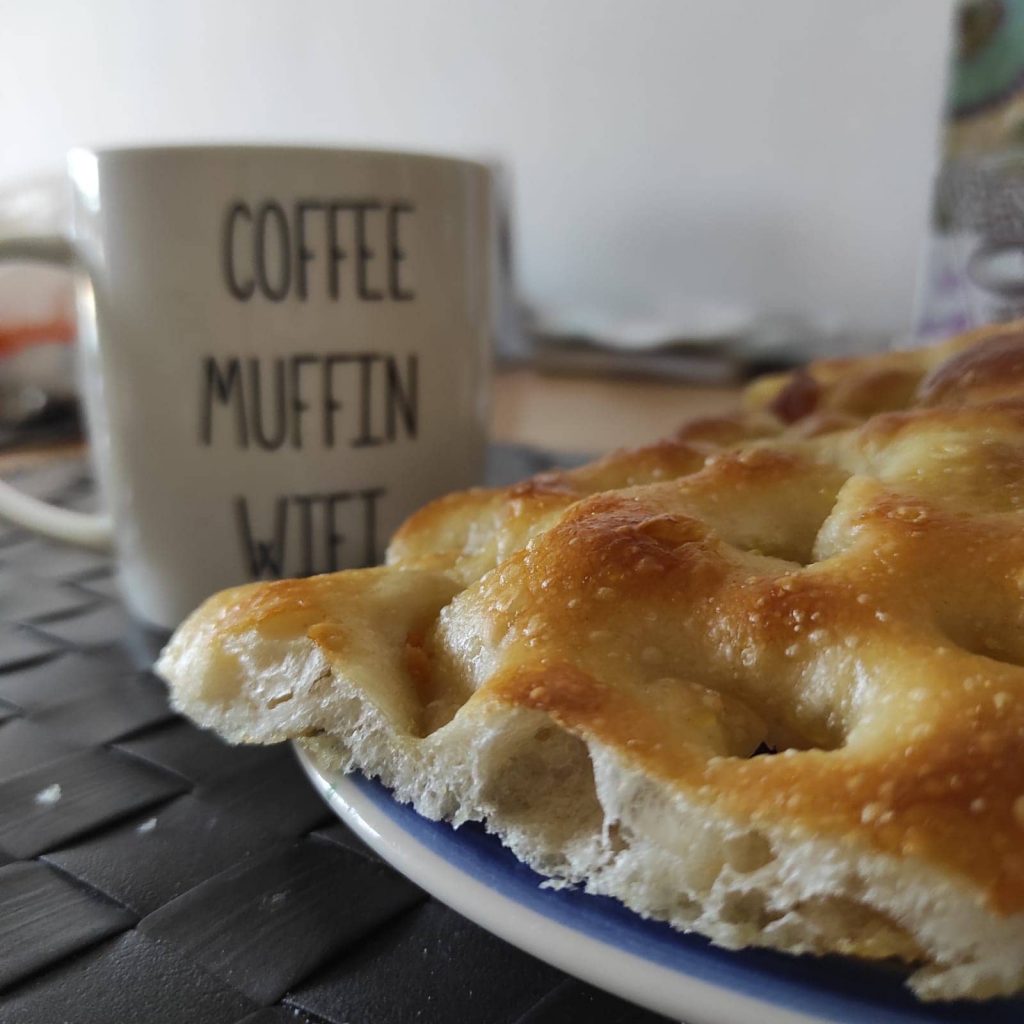 La focaccia genovese è appoggiata su di un piatto vicino alla tazza del caffè latte, pronta per essere mangiata a colazione.