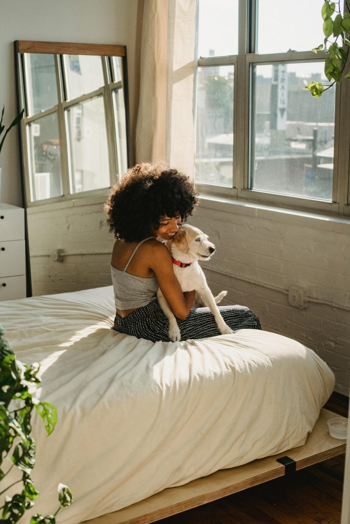 A woman with afro hair lovingly holds a dog in a sunlit bedroom, symbolizing pet companionship.