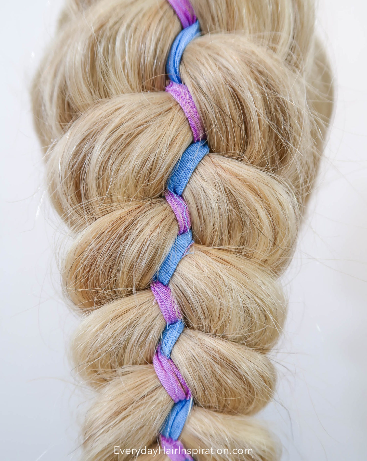 Close up of a blonde 5 strand ribbon braid, with purple and blue colored ribbon zig zagging down the middle of the braid. 