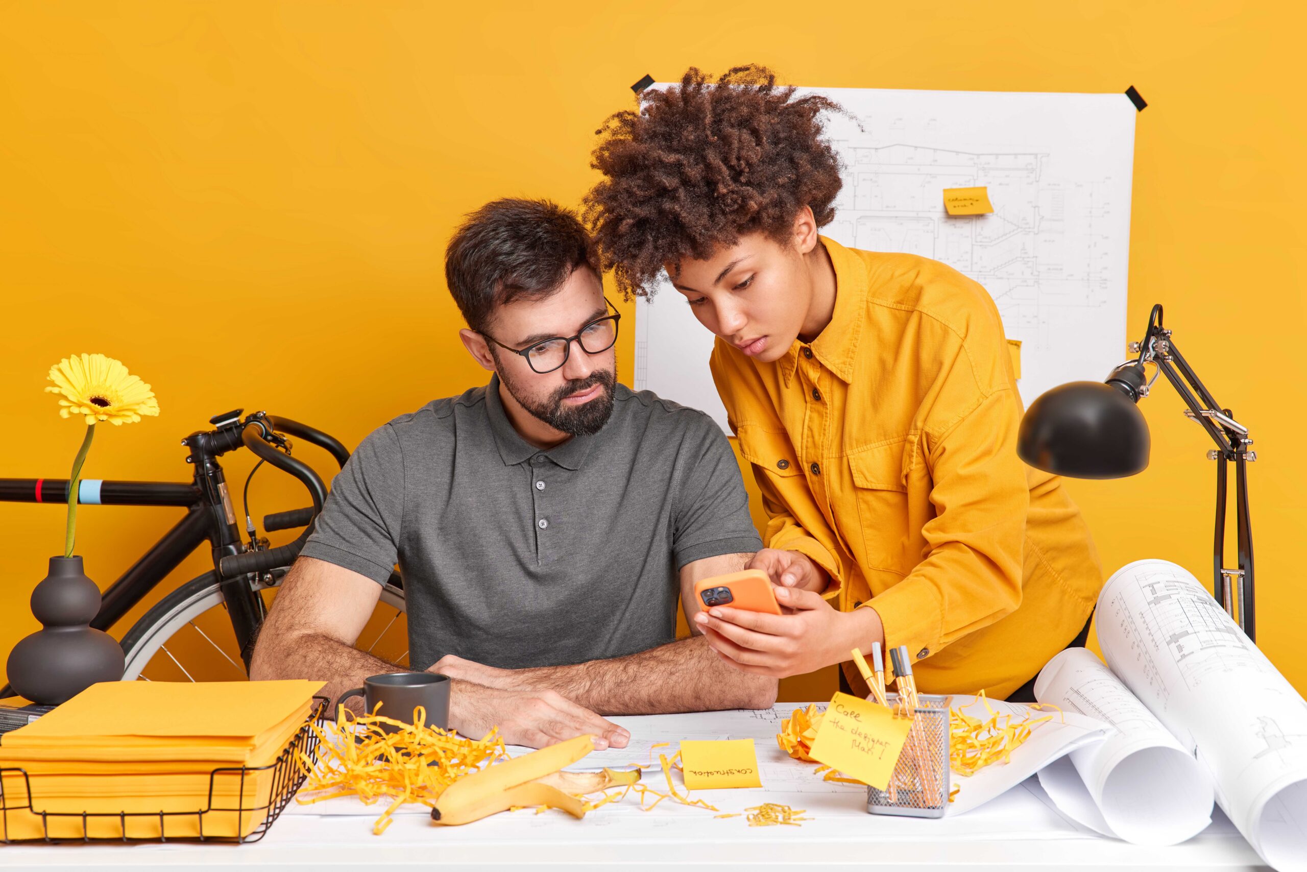 two diverse members of working team think over cretive ideas for future project woman shows design plan in smartphone on webpage pose at desktop isolated over yellow background have informal meeting