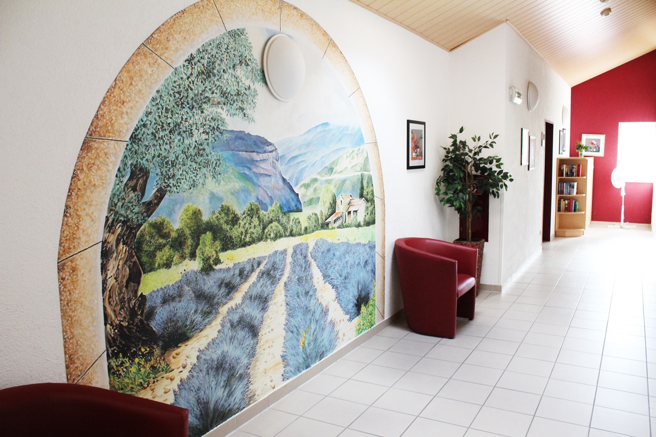 Ein Flurbereich mit heller Wandgestaltung, einem roten Sessel und einer Pflanze. An der Wand befindet sich ein großflächiges Gemälde mit einer Natur- und Landschaftsdarstellung.