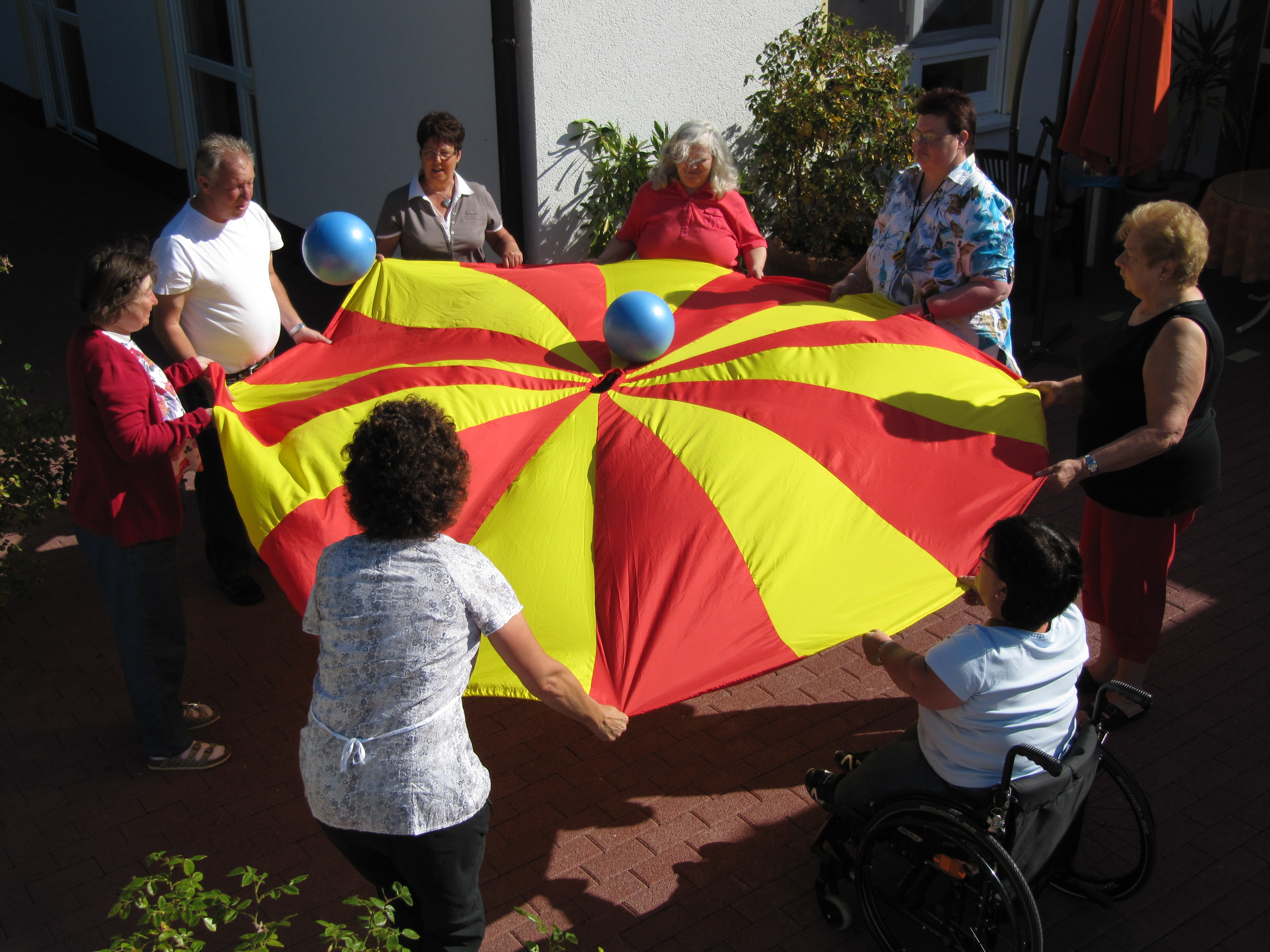 Eine Gruppe von Personen beteiligt sich im Freien an einer gemeinsamen Spielaktivität. Sie halten ein großes rot-gelbes Schwungtuch, auf dem blaue Bälle hin- und herbewegt werden.