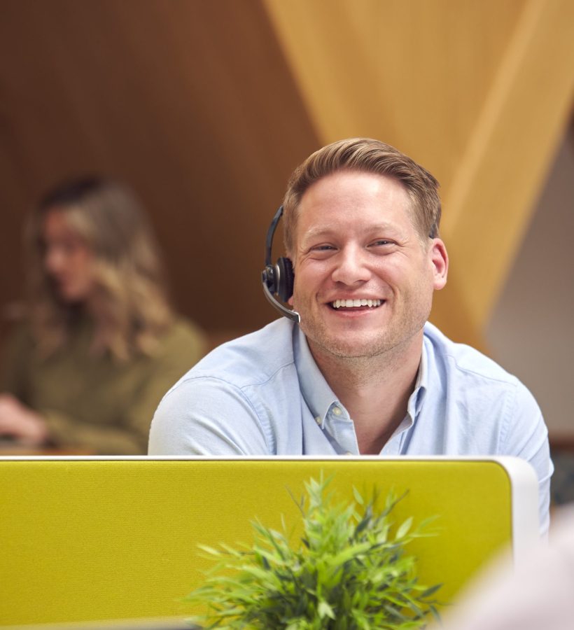 Businessman In Cubicle Wearing Headset Talking To Colleague In Busy Customer Services Centre