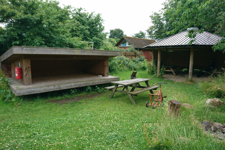 Домик-шелтер слева и крытый павильон со столами и скамейками слева