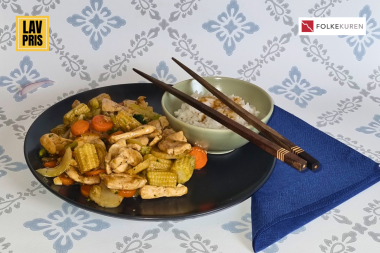 Plate of chicken and vegetable stir-fry (carrots, corn, onions) with a small bowl of rice and pair of chopsticks resting on the bowl, on a blue napkin and decorative backdrop; logos in corners.