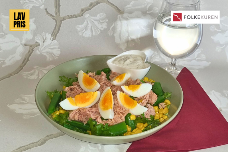 Bowl of tuna salad topped with sliced hard-boiled eggs arranged like petals, with corn and greens; sauce dip on the side and a glass of white wine in the background, plus brand logos in the corners.