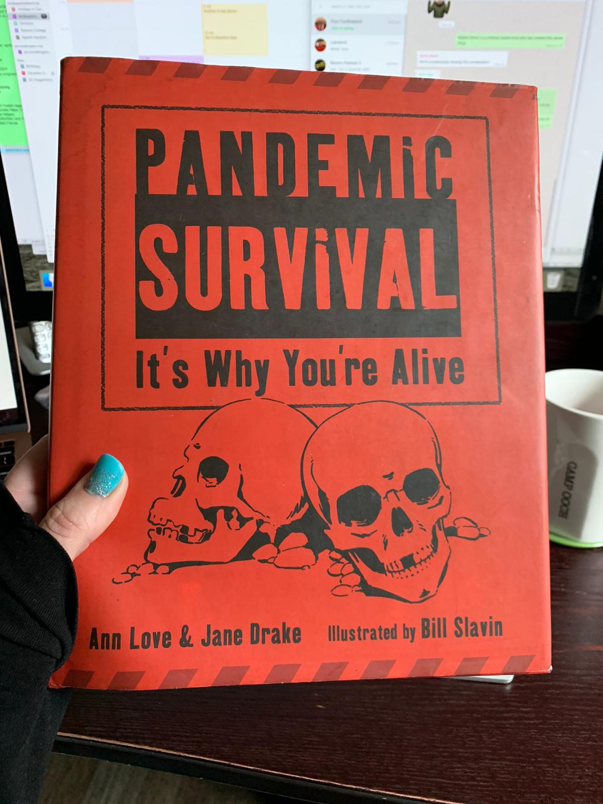 A hand with blue nail polish holding a book. The cover is an alarming shade of red, with two drawn skulls on it. The book title is Pandemic survival - it's why you are alive. Authors Ann Love and Jane Drake, Illustrated by Bill Slavin. 