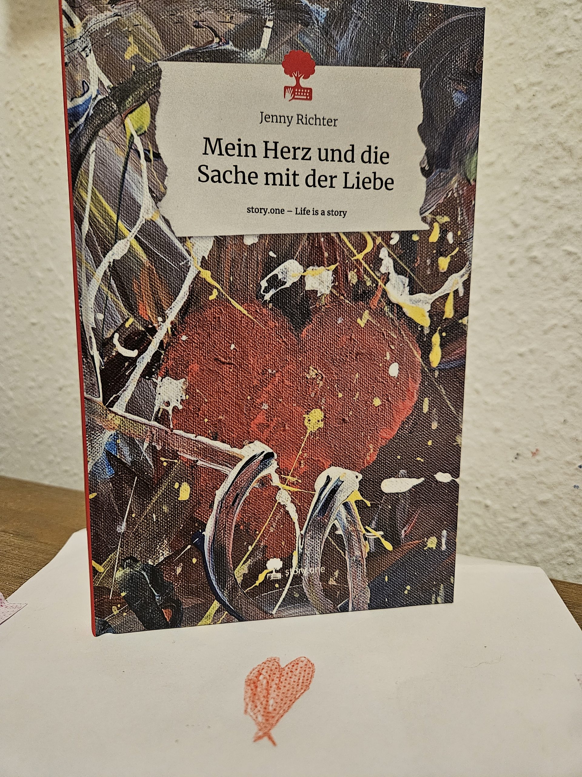 Ein Buch mit einem gemalten Herz und dem Titel Mein Herz und die Sache mit der Realität steht auf einer Unterlage, auf die ein rotes Herz gemalt ist.