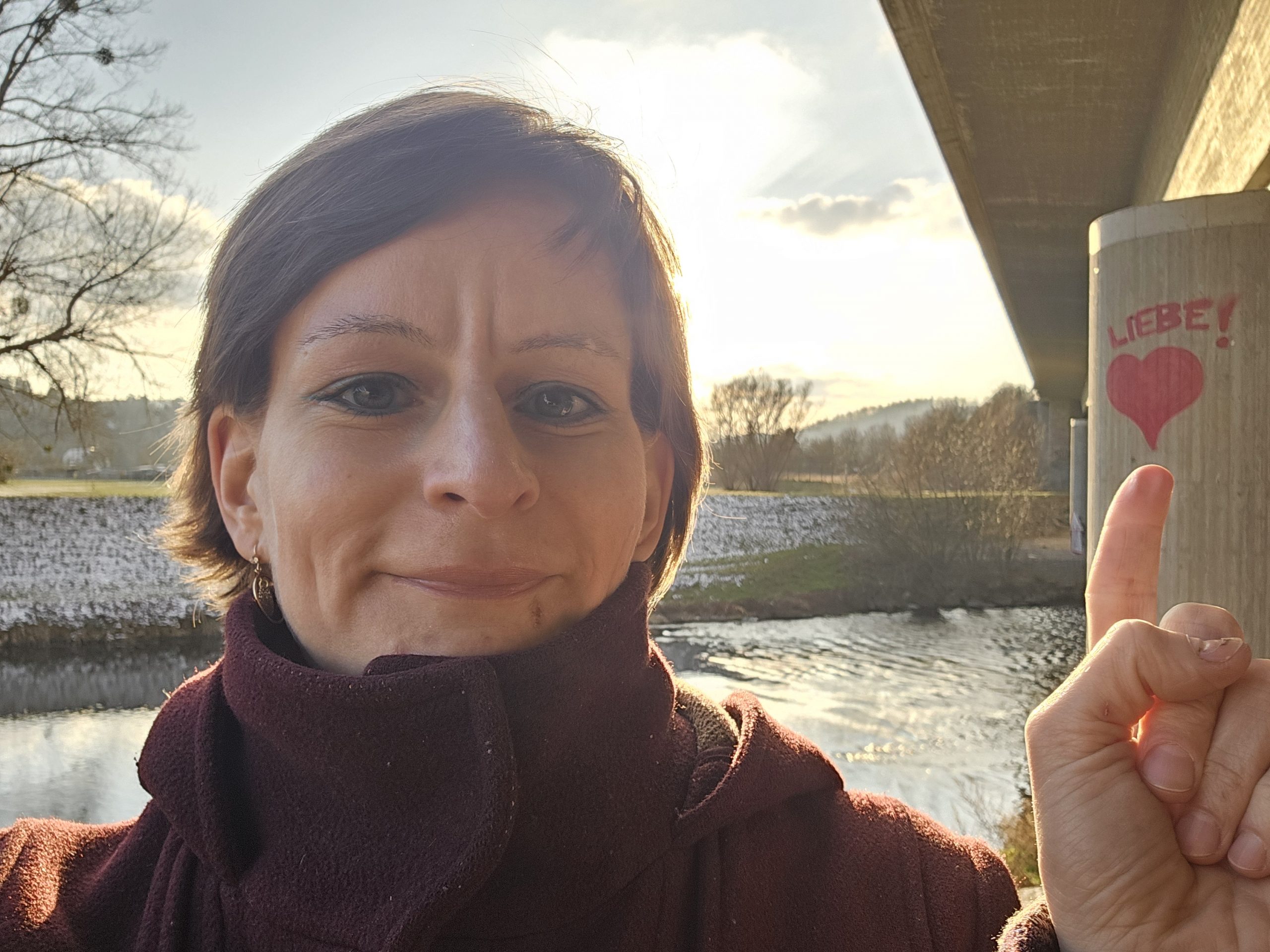 Elsterwanderung Liebe Eine Frau mit kurzen Haaren, die lächelt. Sie steht vor einem Gewässser und zeigt mit dem Finger auf ein an einen Pfeiler gemaltes Herz, über dem steht: Liebe!