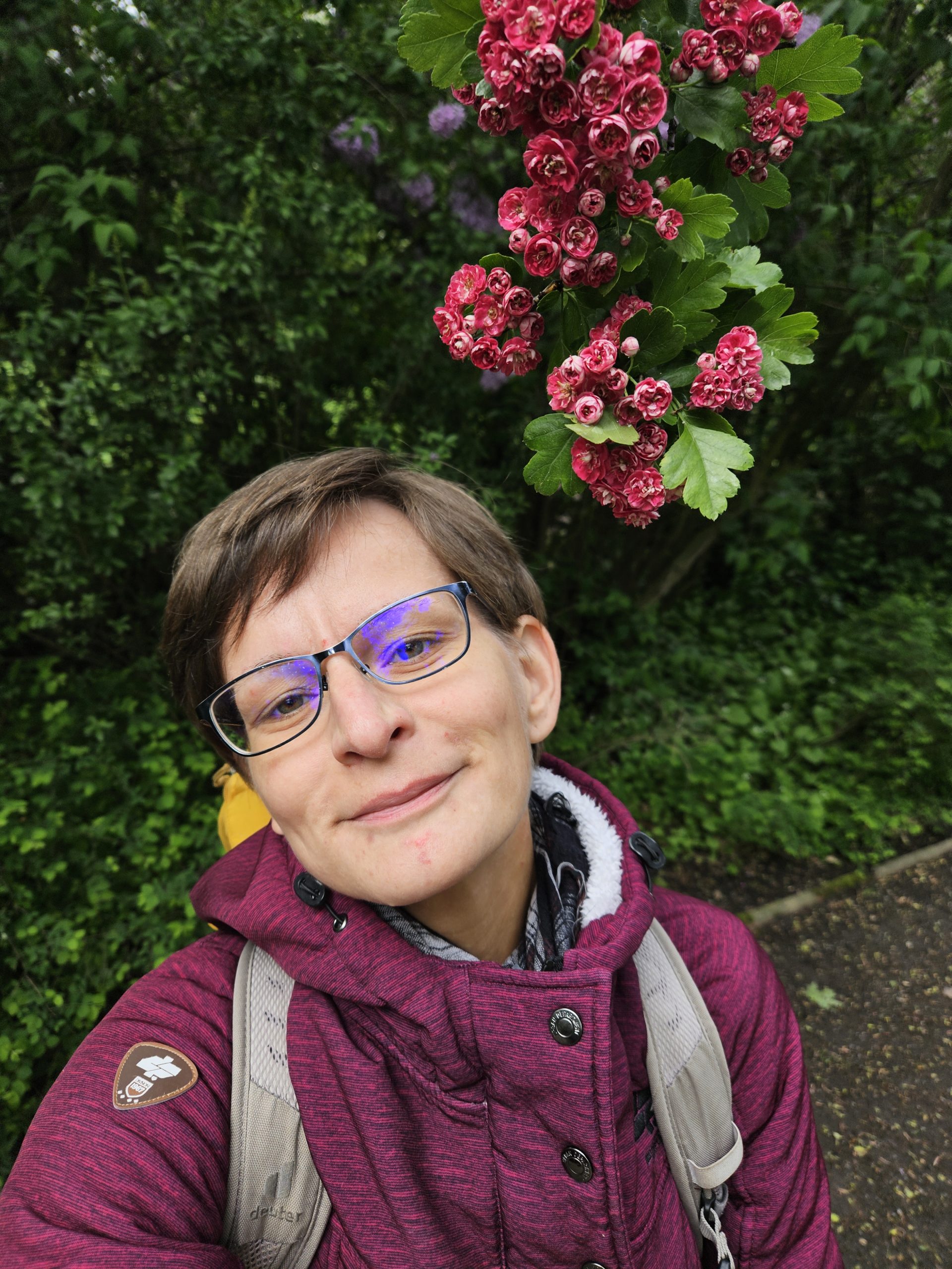 Ich vor Blumen Leipzig Park schräg Eine Frau mit Brille und bordeauxfarbener Jacke links im Bild. Rechts im Bild hängen bordeauxfarbene Blumen von oben herab. Im Hintergrund grüne Büsche und Blätter.