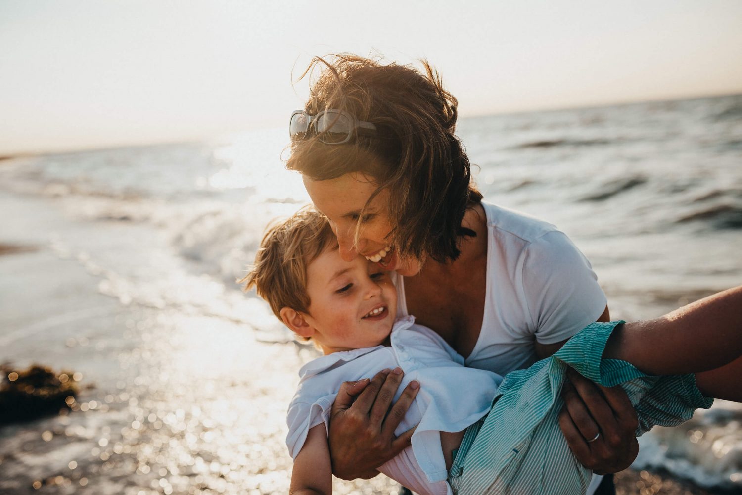 Een mama draagt haar zoontje al lachend uit de zee bij een laagstaande zon