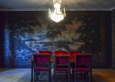 A picture of Italiarummet at Gathenhielmska Huset. Six red chairs, a table and a chandelier. The tapestries on the walls are from Northern Italy and depict an island scene in blues and greens.