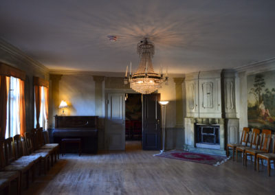 Salongen at Gathenhielmska Huset, with a chandelier, a piano, two lamps, chairs and tapestries on the wall.