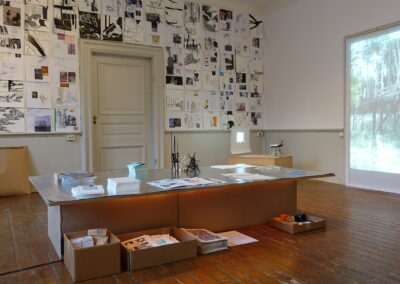 An exhibition in a room, featuring a selection of A3 prints on a wall, a table laid out with materials and two projector screens showing images