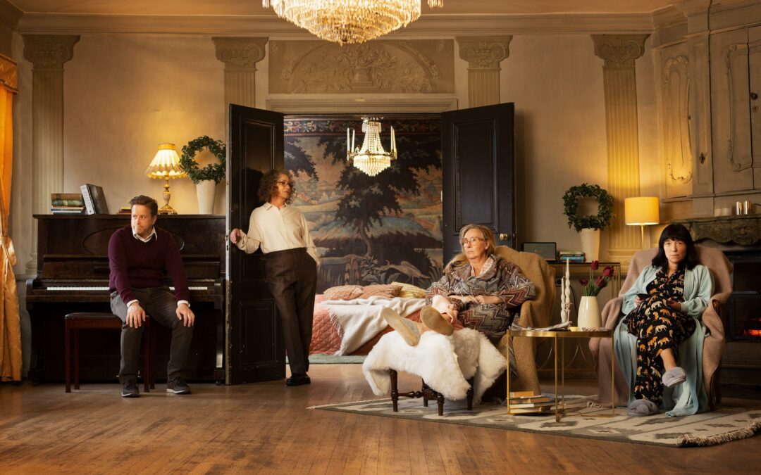 Four actors in a room, one in smart attire sat by a piano, one in smart attire leaning against a door, two in bedclothes sat in armchairs