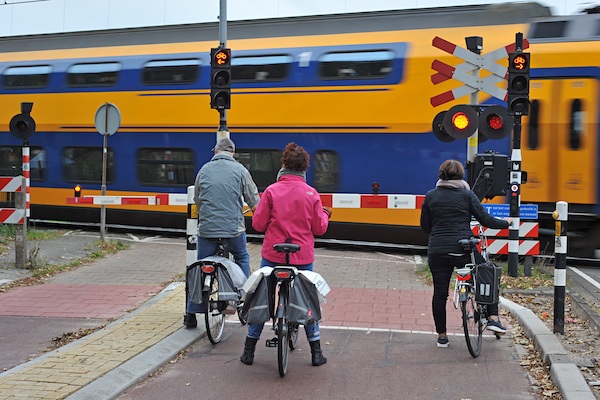 Afbeelding van drie fietser die wachten bij een spoorwegovergang waar trein voorbij rijdt.