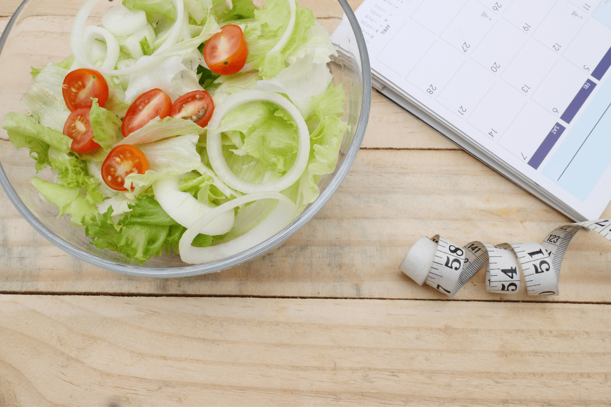 Ein frischer Salat aus Eisbergsalat, Tomaten und Zwiebeln, daneben ein Maßband und ein Kalender. 