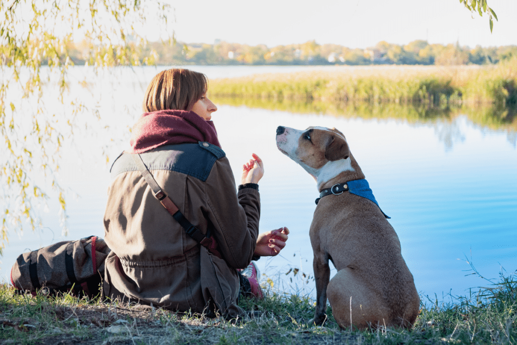 Frau sitzt mit ihrem Hund am See.