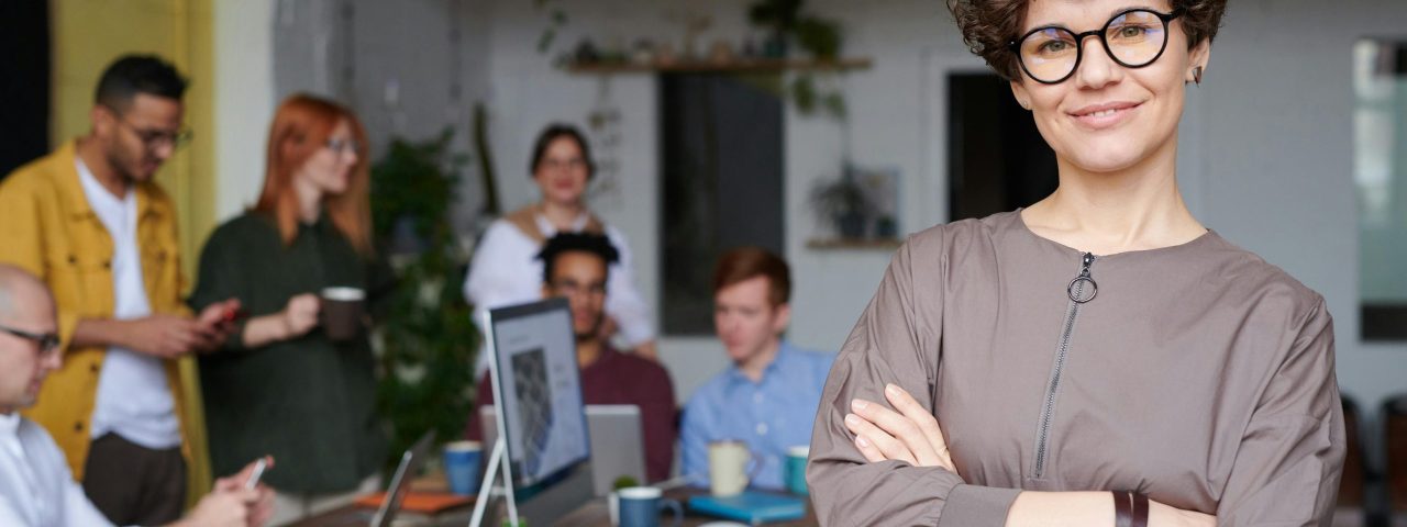 Smiling businesswoman with curly hair stands confidently in a modern office space with colleagues.