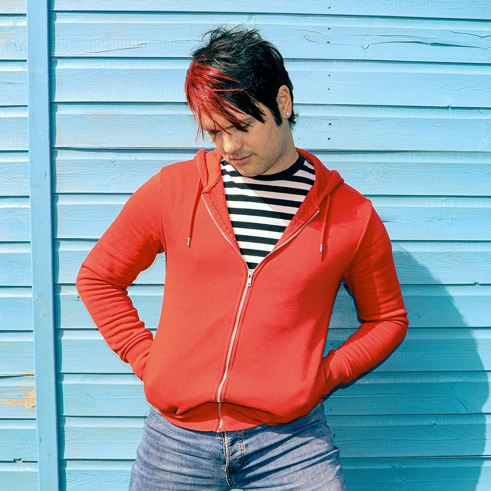 Photo of man in front of light blue shed. Man has black hair with red stripe and is wearing a black and white striped top. He has a red zip up jacket over the top and pale blue jeans.