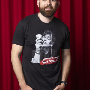 Photo of man against red curtain background. Man has light brown hair . He is wearing a Black t-shirt with a black and white photo of the top-half of a man. Hr is holding a wine glass up against the side of his face. In the bottom right of the photo are the words 'Nick Capaldi' written in white with a red shadow.