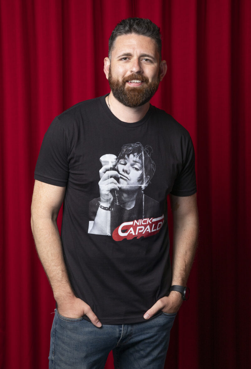 Photo of man against red curtain background. Man has light brown hair . He is wearing a Black t-shirt with a black and white photo of the top-half of a man. Hr is holding a wine glass up against the side of his face. In the bottom right of the photo are the words 'Nick Capaldi' written in white with a red shadow.