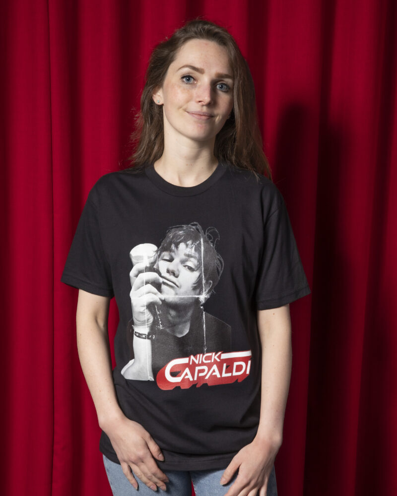 Photo of girl against red curtain background. Girl has long light brown hair with natural curl at end. She is wearing a Black t-shirt with a black and white photo of the top-half of a man. Hr is holding a wine glass up against the side of his face. In the bottom right of the photo are the words 'Nick Capaldi' written in white with a red shadow.