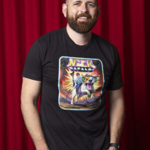Photo of man against red curtain background. Man has light brown hair. He is wearing a Black t-shirt featuring a comic book design of a man wearing a light denim jacket and dark denim jeans with a blue top holding a V-shaped black and pink guitar. He is riding on the back of a giant dog who is coming out of an explosion. The dog is white with an orange patch over his left eye and a purple patch over his right. Written above them both are the words 'Nick Capaldi' Nick is written in yellow with a purple shadow and Capaldi is in electric blue font with a black background. The picture is framed by a yellow inner border and light blue outer border