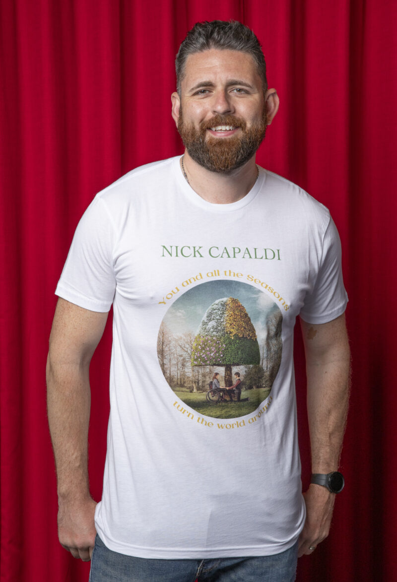 Photo of man against red curtain background. Man has light brown hair. He is wearing a white t-shirt with circular photo on of a woman in a wheelchair and a man kneeling down in front of her. They are holding hands in front of a triangular shaped tree on a Spring day. Arching above the picture are the words 'You and all the Seasons' and curving underneath the picture are the words'turn the world around' both written in gold. In a straight line in green above the gold text are the words 'Nick Capaldi'