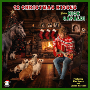 Image shows a man in a cosy cabin front room sitting on an armchair in front of a roaring fire. He is wearing a red and white christmas jumper and is surrounded by 4 dogs, one donkey, a rabbit and a squirrel. He is holding a guitar as if singing to them. In the background, there is a Christmas tree.