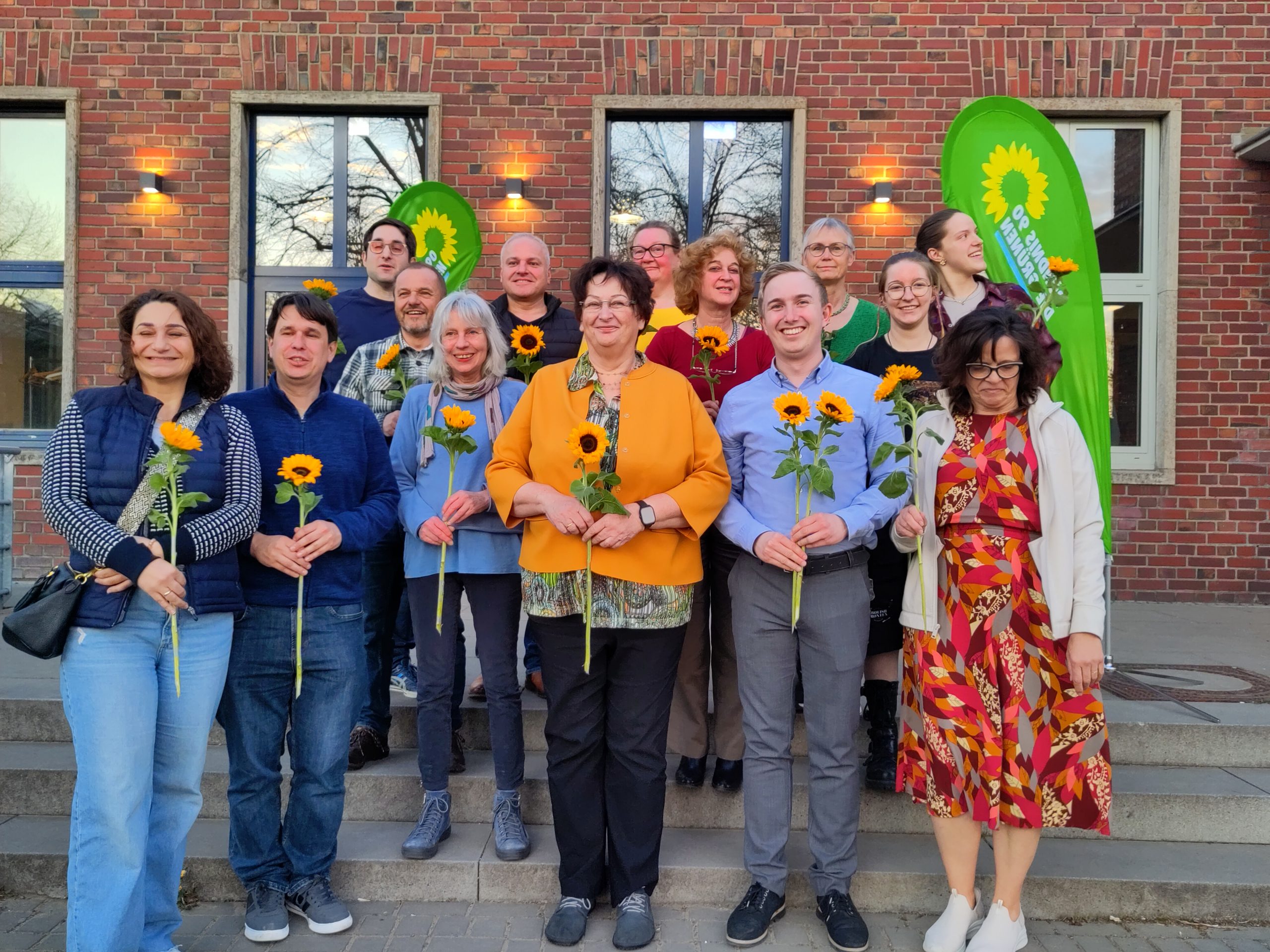 Das Foto zeigt 14 Personen vor dem Kuba Jülich. Alle 14 wurden an diesem Tag als Kandidat*innen für die Kreistagswahl 2025 gewählt. Sie halten Sonnenblumen in der Hand und lächeln in die Kamera. Hinter ihnen sind noch zwei Beachflags mit BÜNDNIS 90/Die Grünen Logo.