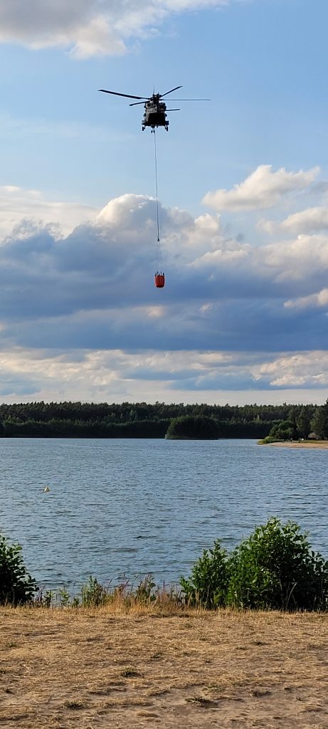 Hubschrauber mit angebrachten Behälter fliegt über See um ihn mit Wasser zu füllen für die Löscharbeiten