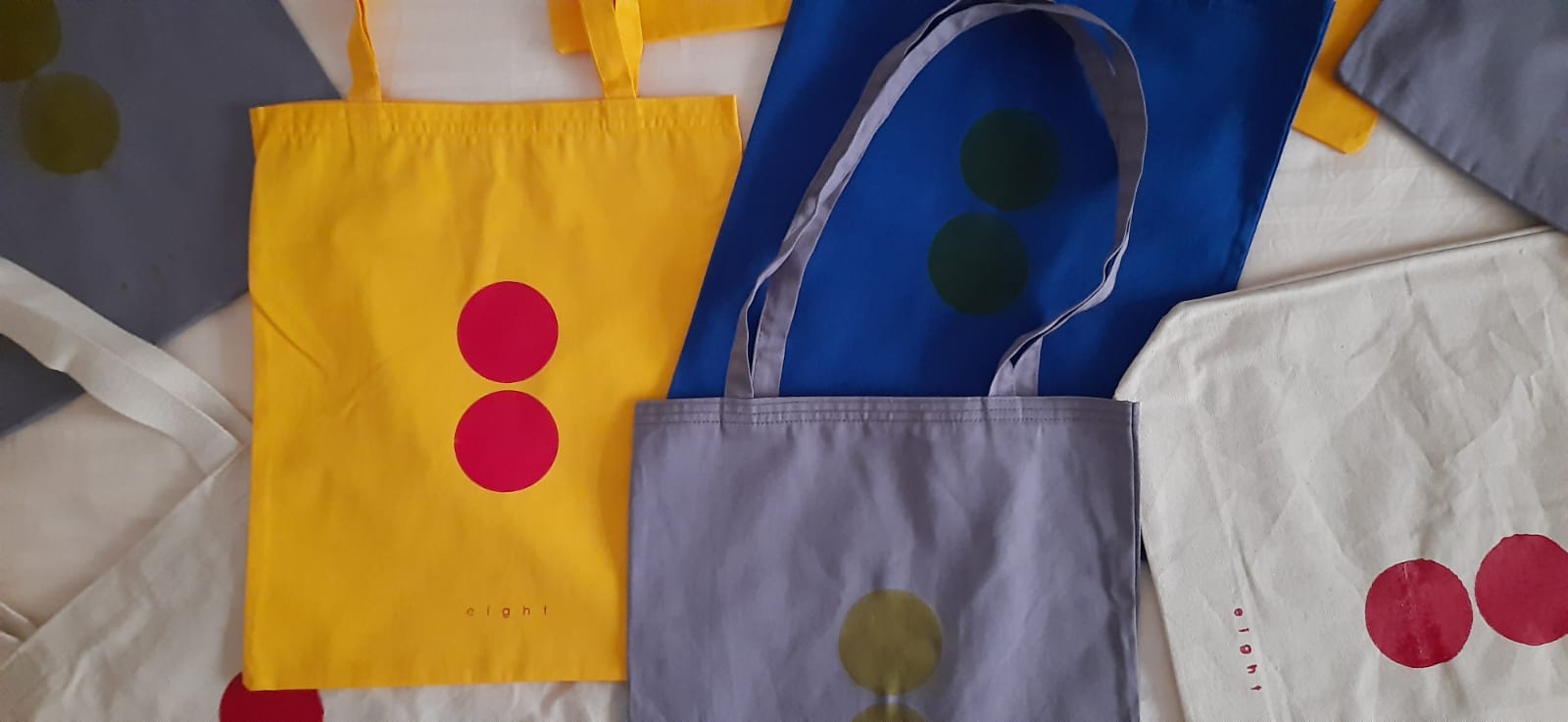 Eight totebags of a variety of colors are spread around overlapping one another.