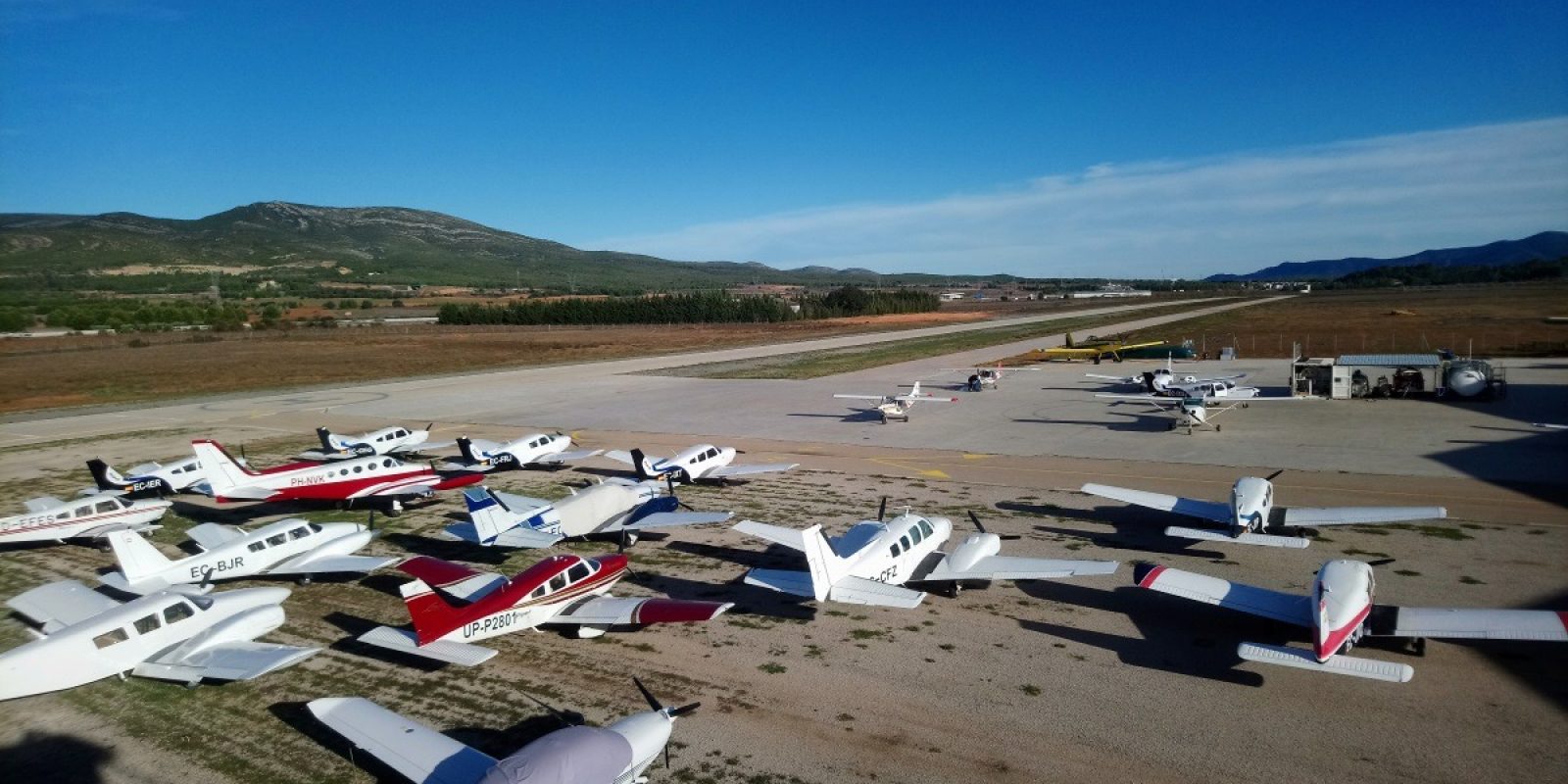 Een rijtje GA-toestellen broederlijk naast elkaar op het vliegveld van Requena - El Rebollar (LERE). Op de achtergrond het geelgeschilderde blusvliegtuig van het lokale brandweerkorps. (Foto Thomas Cruysberghs)