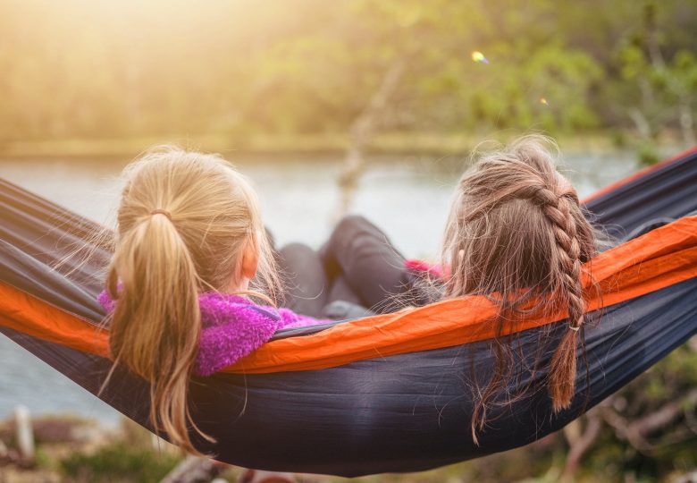 Två flickors bakhuvud i hängmatta. Vatten och skog framför dem. Fläta och hästsvans i håret.