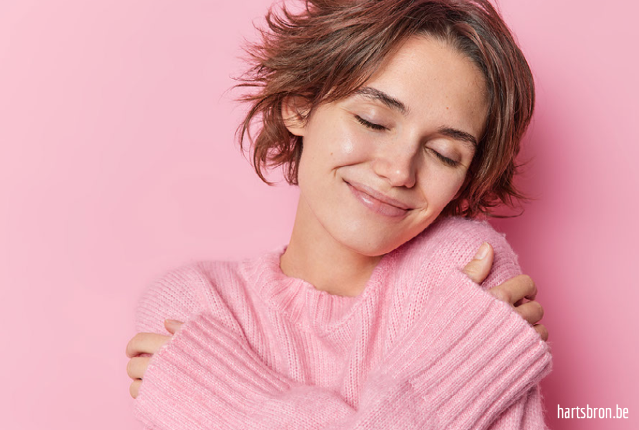 glimlachende jonge vrouw omarmt zichzelf met de ogen gesloten, zachte roze trui