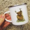 A hand is holding the Heelan Coo Enamel Mug, featuring a charming illustration of a shaggy Highland cow adorned with birds perched on its horns. This whimsical Scottish design is set against a backdrop of lush green grass and surrounded by an eye-catching metallic rim. The textured beige rug in the background adds warmth and enhances this delightful scene, making it perfect for showcasing rustic charm kitchenware or Scottish-themed gifts.
